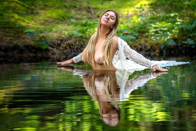 Frau mit weißer Spitze im Wasser, ihre Spiegelung zwischen grünen Uferpflanzen.